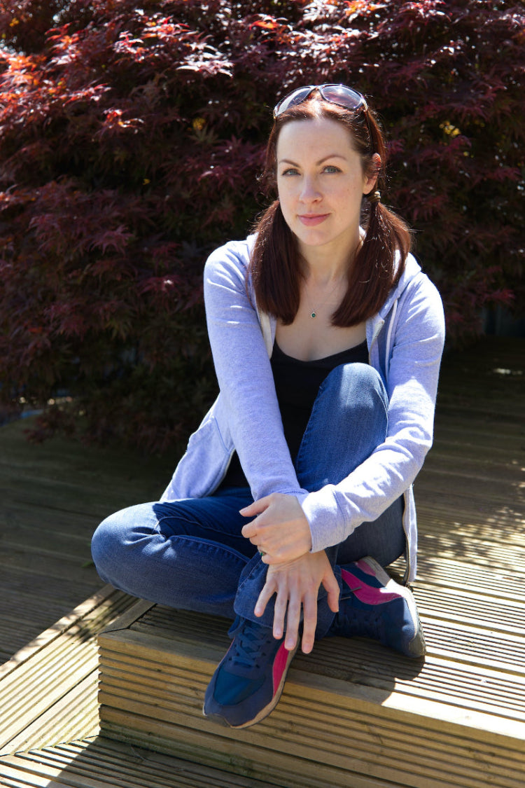 Emma Gorton-Garlick smiling at the camera, sitting outside in front of a lush acer, enjoying the natural surroundings that inspire her designs.