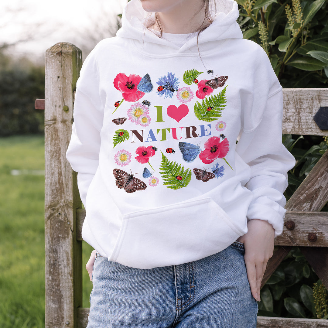 Teenage girl wearing a white hoodie with "I Love Nature" design and jeans, standing against a wooden gate with field and greenery in background.