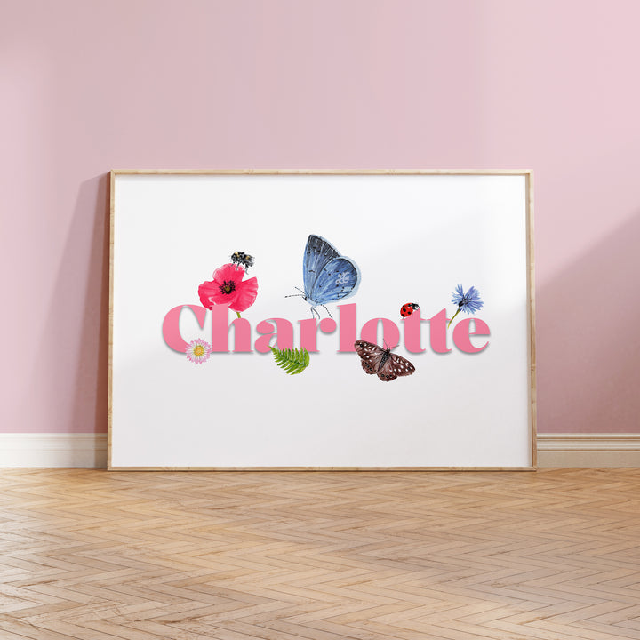 Personalised wildlife meadow name print with "Charlotte" in soft pink, framed on a wooden floor against a painted pink wall, featuring UK meadow flowers and insects.