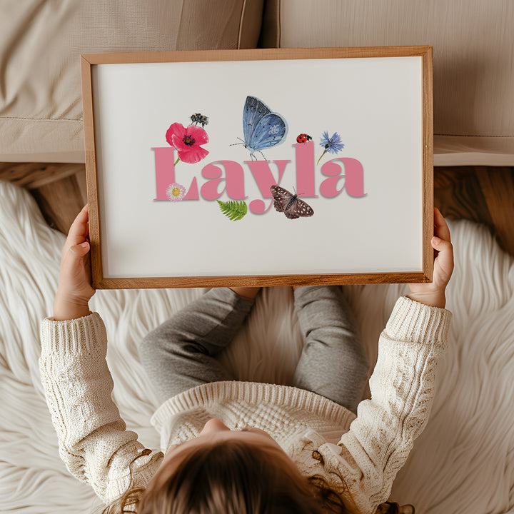 Child holding a framed personalised wildlife meadow name print with "Layla" in soft pink,  featuring UK meadow flowers and insects.