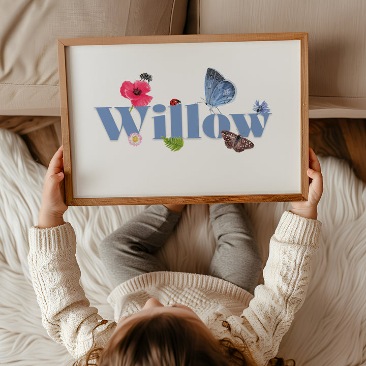 Child holding a framed personalised wildlife meadow name print with "Willow" in soft blue,  featuring UK meadow flowers and insects.