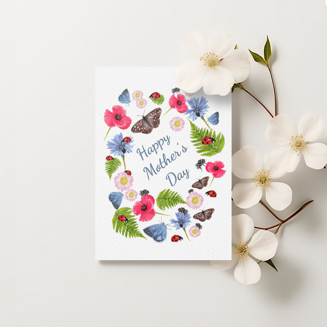 Happy Mother’s Day card with a border of watercolour poppies, butterflies, bees, and daisies, printed on textured card with flowers beside it.