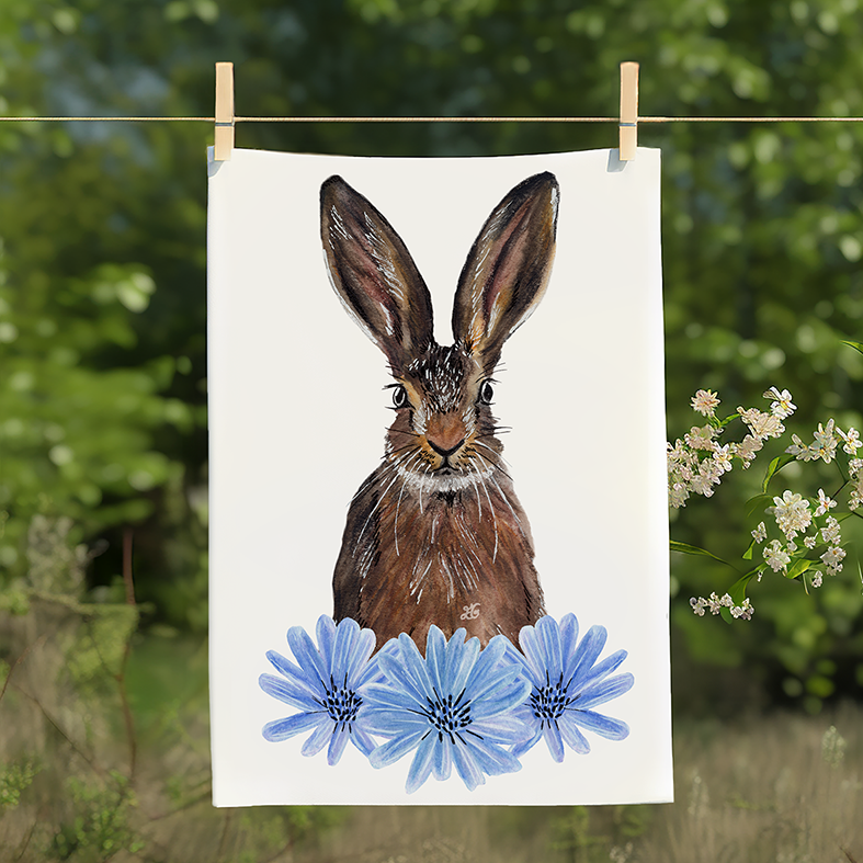 Illustrated tea towel featuring a woodland hare nestled in chicory flowers, designed by Emma Gorton-Garlick, hanging on a washing line in a garden setting with greenery and blossom in the background.