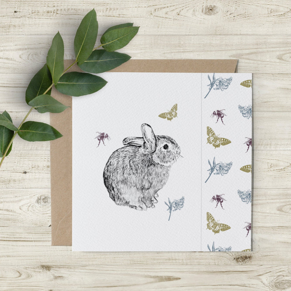 Bunny and Butterfly card outer design, featuring a charcoal pencil sketch of Bryn the rabbit, with coloured butterflies, flowers, and bees, and a cutaway edge design, eco-friendly and made in the UK by artist Emma Gorton