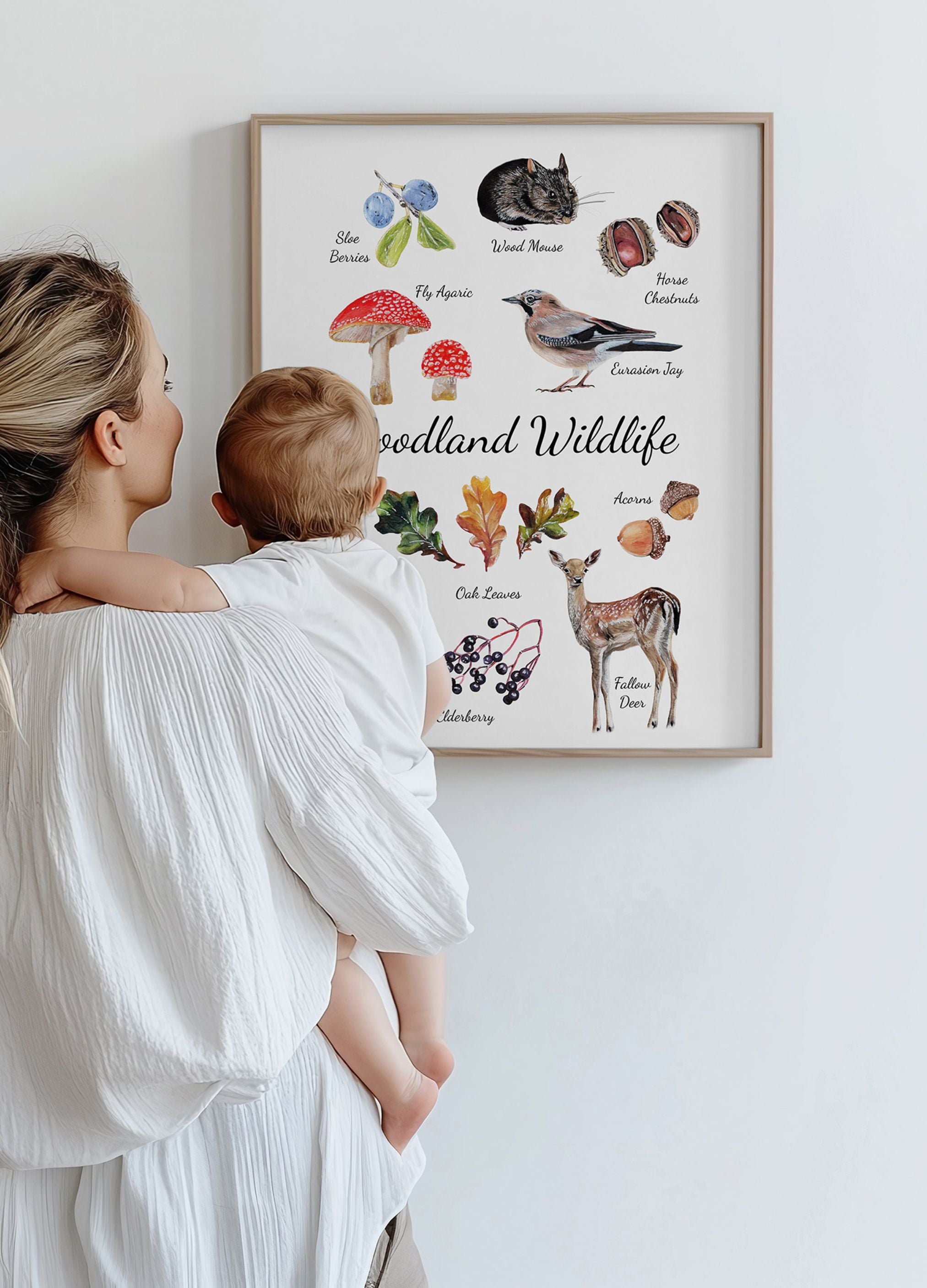 Woman holding a child in front of a framed illustration of woodland wildlife on a white wall.