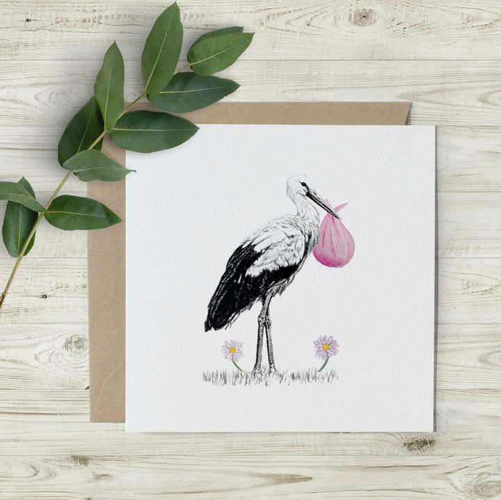 New baby girl card featuring a hand-drawn stork carrying a pink cloth bundle, printed on eco-friendly textured paper. A percentage of sales supports wildlife charities.