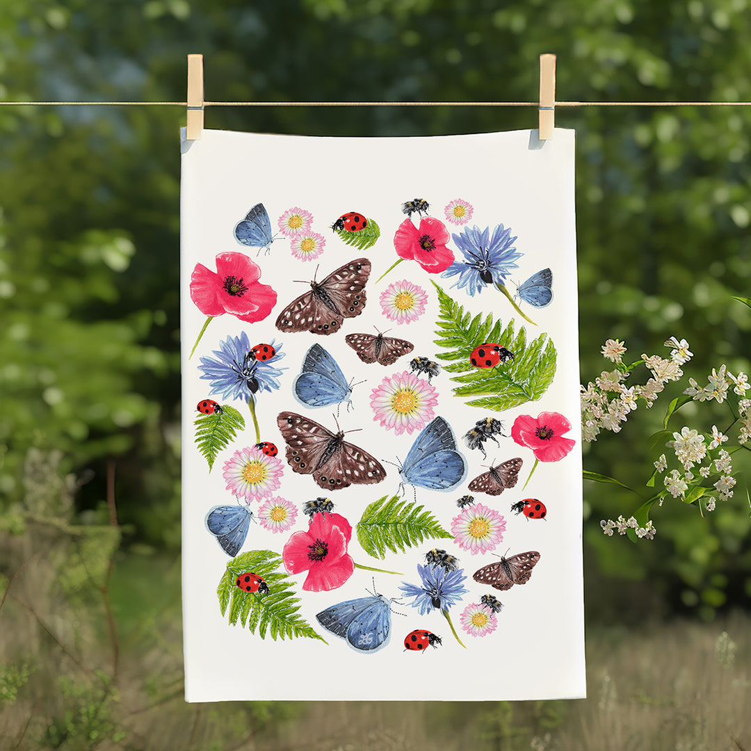 Summer Meadow cotton tea towel by Mummy and Bear hanging on a washing line outdoors, with greenery and flowers in the background – featuring bees, butterflies and wildflowers