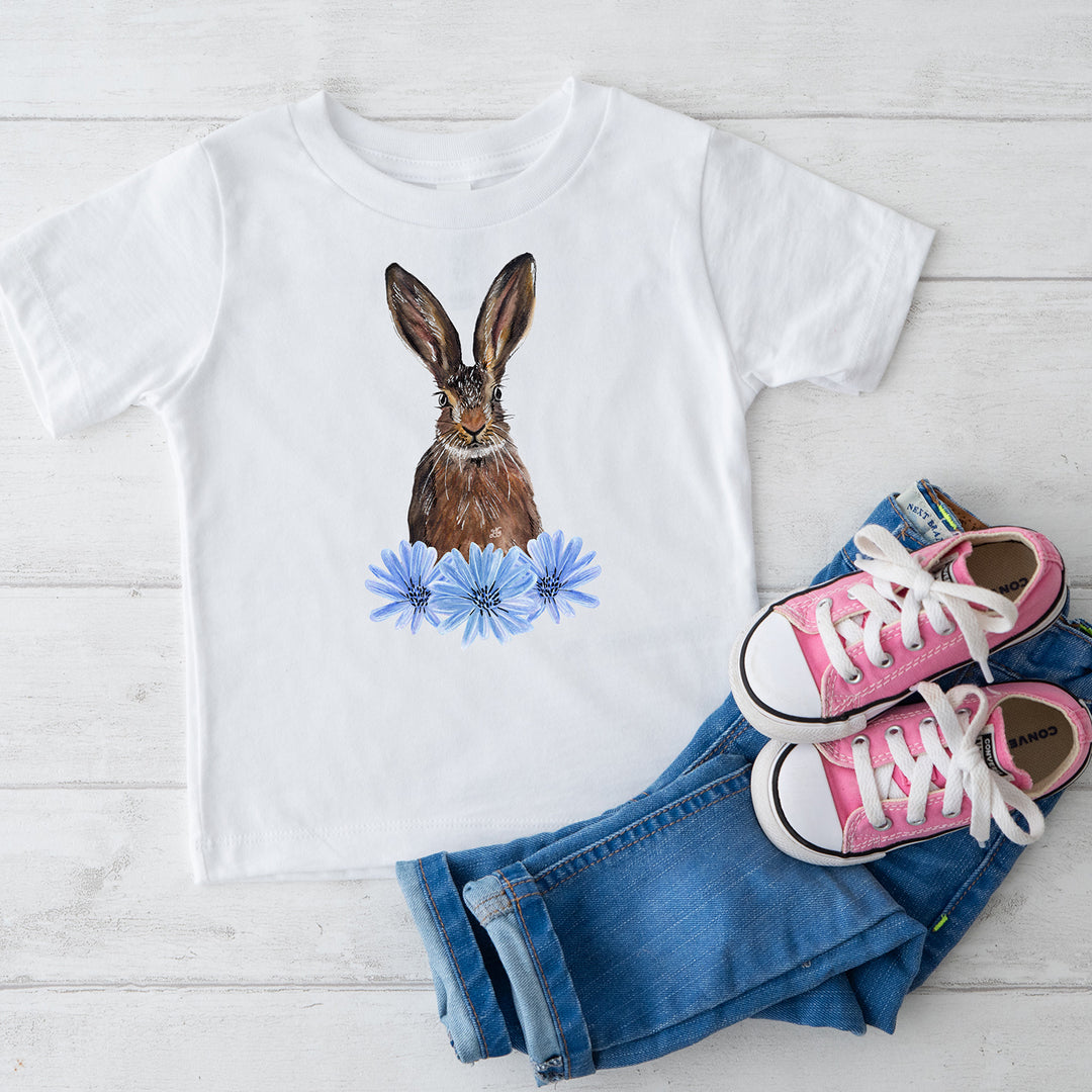 Flatlay image with white tshirt with a hare and flower design, pictured on a pale wooden background with some jeans and pink trainers