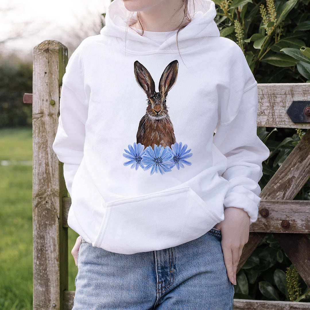 Older girl wearing a white hoodie featuring a woodland hare and chicory flower design by Emma Gorton-Garlick, standing in front of a wooden gate in the countryside.